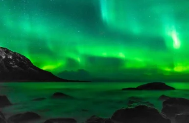 Green aurora borealis over rocky shoreline at night.